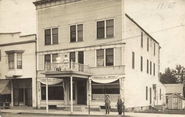 Photographic postcard of the exterior of the three-story Commercial Hotel. The building has a false front. Two women stand on the balcony and two men stand near a hand-pump at the curb. A glider swing is in the yard on the right. Another building on the left has a sign that reads "Crystal."