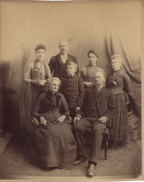 Full-length studio portrait of the Gunder Larson Family. Front row: Mary Jane Rodgers Larson, Arthur Larson, Gunder Larson. Back row: Margaret Slossen Larson, Sam Larson, Adina Larson, Florence Larson. The background appears to be a painted backdrop, with a curtain on one side.