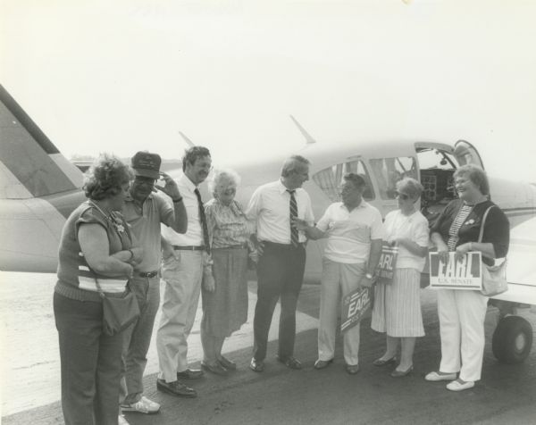 Former Wisconsin Governor Anthony Earl stands outdoors near an airplane with a group of supporters. He made an unsuccessful run for the United States Senate, and lost in the primary to Herb Kohl who went on to win the general election. Of Earl's supporters in the photograph, only Congressman David Obey (third from the left) has been identified.