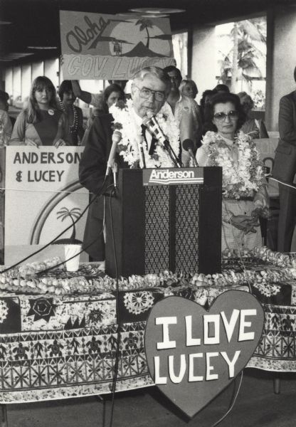 Former Wisconsin Governor Patrick J. Lucey, together with Mrs. Lucey, campaigning for vice president on John Anderson's National Unity ticket. They are both wearing leis. A sign below reads, "I Love Lucey."