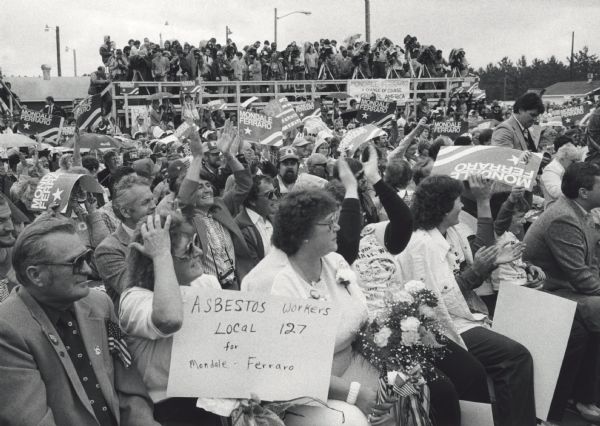 The audience at the Lincoln County fairgrounds who gathered to hear Democratic candidates Walter Mondale and Geraldine Ferraro, with the national media covering the candidates on the platform behind them. The hand-lettered sign a woman holds in the foreground reads, "Asbestos Workers Local 127 for Mondale-Ferraro." A woman next to her holds a bouquet of flowers and miniature American flags.