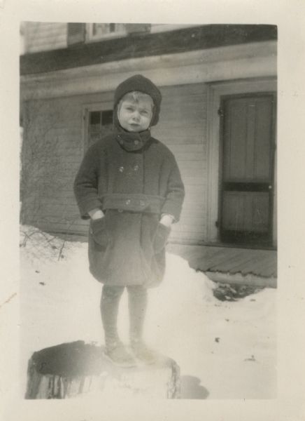 Dorothy Sunne (b. 1922) stands on a stump in front of her house. She is wearing a winter coat with a cap, stockings and shoes.