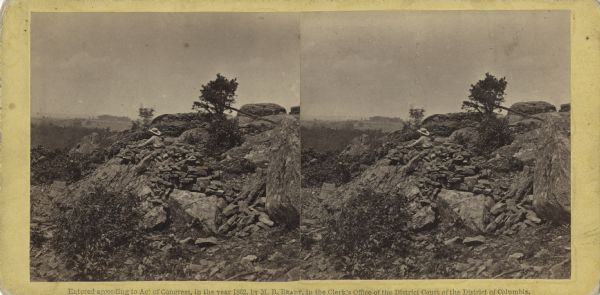 Stereograph of breast works on a rocky outcropping on the site of the Battle of Gettysburg. A man can be seen standing behind the breast works. He is gazing into the distance. On the reverse, written in pencil is "On the left of our line."