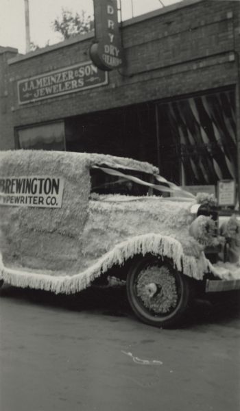 Madison businesses had a parade to encourage customers to shop. This is the Brewington Typewriter Company entry. It says Brewington Typewriter Company on the sides and Royal on the back. They are parked in front of 3F Dry Cleaners and J.A. Meinzer & Son Jewelers.