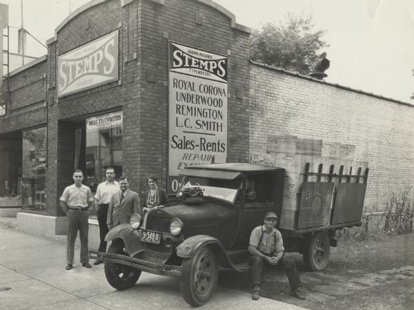 Stanley P. Stemp (owner), third from the left, and three employees stand on the sidewalk next to an Ed Klief Transfer Line delivery truck and driver. Behind them the Stemp Typewriter Company can be seen, located at 533 State Street. The truck is filled with a large delivery of office machines.