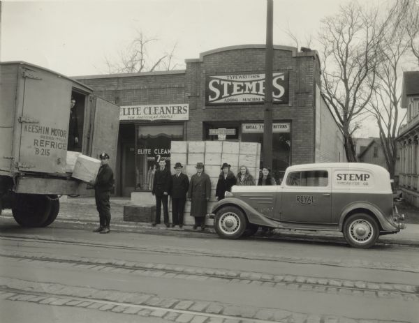 View from across State Street of men and women standing in front of the Stemp Typewriter Company as deliveries are stacked on the sidewalk. Stanley P. Stemp (owner) is fourth from the left and his wife, Elisabeth, is standing next to him. A Stemp delivery truck is parked to the right and a large delivery truck is on the left. A deliveryman is unloading boxes. The business on the left is Elite Cleaners, "For Particular People."