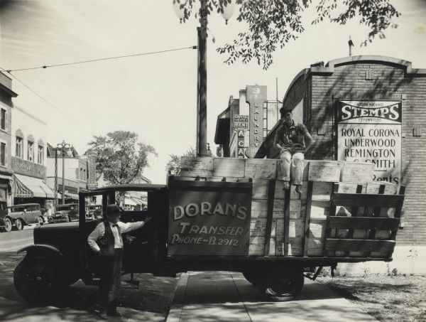 View down the sidewalk of two men standing near a Doran's Tranfer delivery truck next to the Stemp Typewriter Company. One man is standing next to the cab and the other is perched on the load of typewriter boxes. The Stemp Typewriter Company sign can be seen behind the truck box. Other business signs appear in the background, 3F Laundry, Capital City Rent a Car, Chocolate Shop and a Delicatessen.