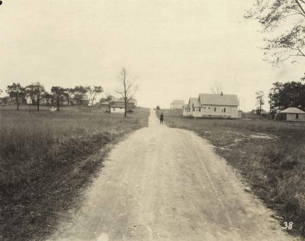 View down dirt road running through homes and trees in a neighborhood. A man stands in the road. Text at foot of photograph reads, "Toelle Ave. 138+12 Looking South from Line."