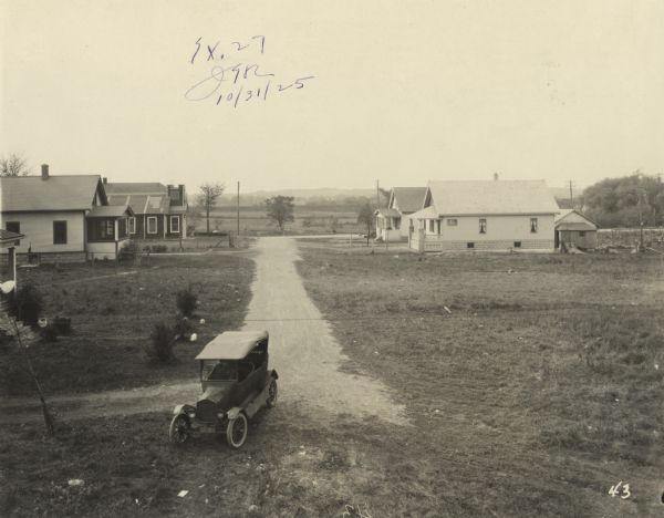 View of dirt road running through homes and trees in a neighborhood. An automobile is parked in the foreground, at the end of the road. Text at foot of photograph reads, "McMyron St. 225+90 Looking West from Line." Handwritten in ink at top, "Ex. 27, (unreadable initials), 10/31/25."