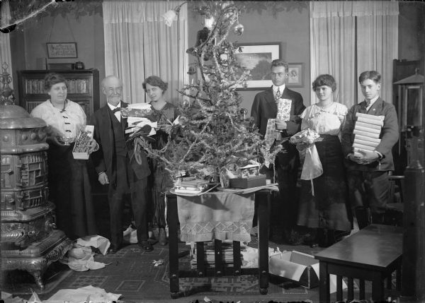 Family holiday scene in a living room or parlor. A Christmas tree is perched on a table in the center of the image. It is trimmed with tinsel garland, shiny balls, paper bells and candles. On the right, two young men and a young woman stand with their arms full of gifts. On the left, two women and a man stand between the tree and a large cast iron stove. They also have gifts in their arms. More gifts and other items are stacked under the tree. Among the gifts are a box of nuts with a  silver dish, leather mittens and slippers, ties, books, picture frames, perfume, and an apron. Torn paper and open boxes litter the floor. A stack of books or magazines appear under the table. Decorations hang from the ceiling light fixture. The men are wearing suits and the women are wearing dresses.