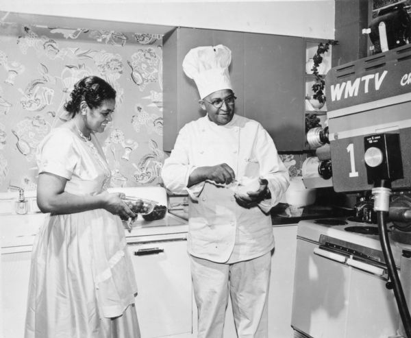 Chef Carson Gulley and Beatrice Gulley, Carson's wife, who appeared in this program with Gulley, on Madison television station WMTV. 

They are being filmed for his weekly television program, "What's Cooking." His television show ran from 1953 until 1962. In 1953 he also hosted a twice weekly cooking program called "WIBA Cooking School Of The Air" that aired on a local radio station. The dining hall where he served as head chef is now known as Carson Gulley Commons.