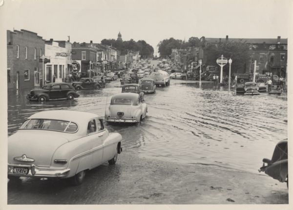 View up hill of cars driving in the flooded street of the downtown area. The downtown had also flooded in 1923 and 1937. The Pecatonica River rose rapidly after two inches of rain began on February 27. Hard frozen ground sent all the water toward the town, where the river rose a foot each hour before cresting at 18 feet. Two blocks of the business district on Main Street fell under several feet of water, the fairgrounds lay under five feet, and the Milwaukee Road discontinued trains for nearly a week.