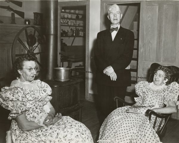 Carl Sandburg posing with two of the guides at Wade house during his visit to the restored stagecoach inn for its dedication as a historic site. Speaking at the dedication ceremonies, Sandburg said, "After all of us here have vanished, other generations will come here and say how lovely, how thoughtful it was of those people way back there to keep this house as it was when it was a living thing." Sandburg is wearing a suit and the guides are wearing period dresses. A woodburning stove, firearms, mantel, and other historic items are displayed behind a chained off area in the background.