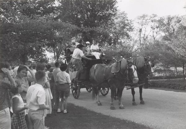 Visitors enjoy a stagecoach ride, an attraction at Old Wade House during the August "twilight tours" of this historic site. A team of two horses stand in the road, waiting to pull the stagecoach as the driver climbs to his seat. Adults and children are seated in the stagecoach as a line of people watches and waits for their turn.