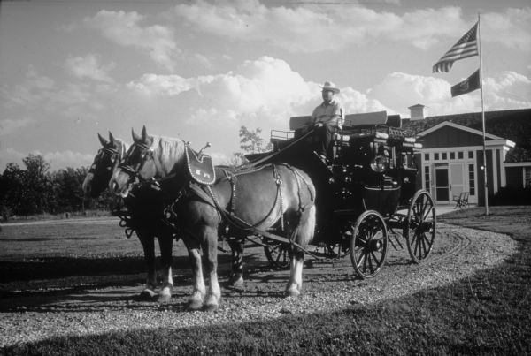 Grand opening of the Wesley W. Jung Carriage Museum. A stagecoach is pulled by horses from the Jung Carriage Museum at Wade House along a curved gravel drive. The driver is seated on top and is wearing a western hat. The Jung Carriage Museum is in the background. A flagpole flies the American and Wisconsin flags.