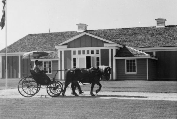 Grand opening of the Wesley W. Jung Carriage Museum. Side view of a carriage with a fringed shade, pulled by a horse, traveling in front of the museum on the grounds of the Old Wade House. A man and woman are passengers, and a man walks on the far side of the horse. A flag on a flagpole is on the far left.
