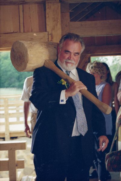 Herbert V. Kohler, Jr. presented with an oversized wooden mallet at the grand opening of the rebuilt Herrling Sawmill. He is wearing a suit with a boutonniere on his lapel and blue cufflinks. A group of people stand behind him. A wooden deck and railings are in the background outside of the building.