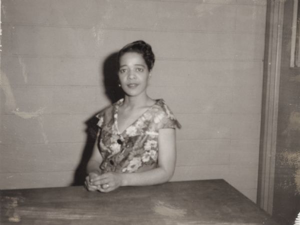 Vel Phillips seated at a table. She is wearing a flowered dress and has her hands clasped together.