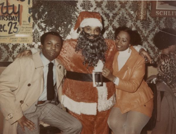 An African American Santa Claus has his arms around an African American man and woman in what appears to be a restaurant. There is wall paper and posters behind them on the wall, and a hanging plant appears on the upper left. Another woman is seated facing away on the right.