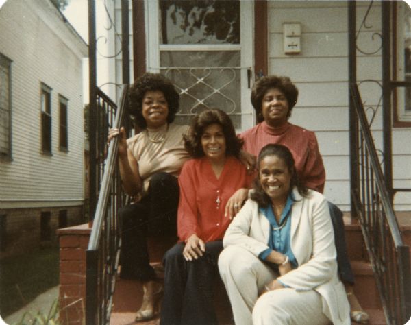 Four African American women posing while sitting on porch steps. Three of them are wearing slacks and blouses, while the woman in front is wearing a white pantsuit. The steps and porch have wrought iron hand rails. The house has white siding and red trim. Another house is on the left.
