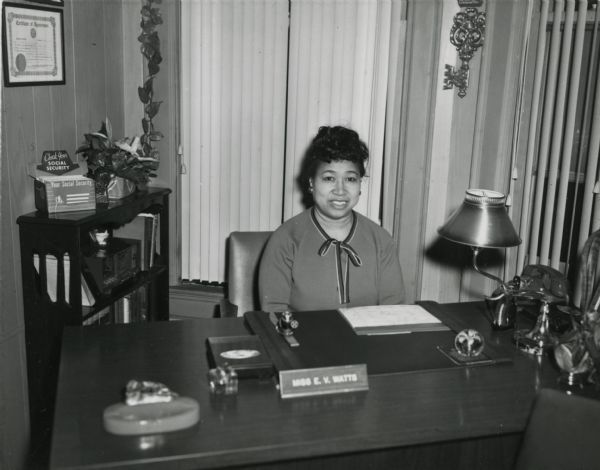 Photograph of Eydie V. Watts, member of the Milwaukee City Civil Service Commssion. Ms. Watts sits at her desk in an office. She is wearing a dress or blouse with a bow at the neck. The usual office equipment is on her desk along with a name plate. In the upper left corner is a Certificate of Appointment, and beneath is a bookcase with books, a radio and a literature box for Social Security flyers.