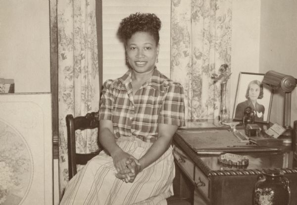 Photograph of Eydie V. Watts, member of the Milwaukee City Civil Service Commission. Ms. Watts sits on a chair with her hands clasped. She is wearing a striped skirt, plaid blouse and earrings. On the desk to her right is a framed portrait of a woman and a flower in a vase. Behind her is a window with the blinds and drapes.
