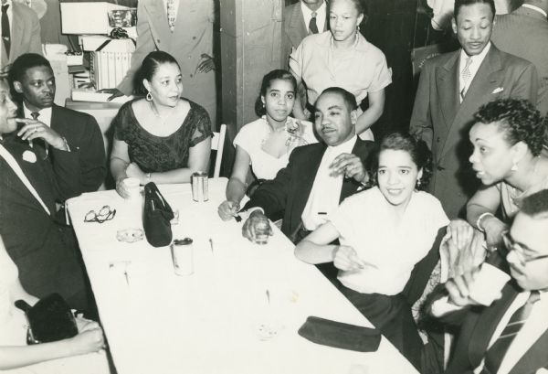 Vel Phillips sits at a table with a group of African American men and women. The table is covered with handbags, eyeglasses, and beverages. Several more people are standing in the background, two inside an office door frame. The women are wearing dresses and jewelry and the men are wearing suits and ties.