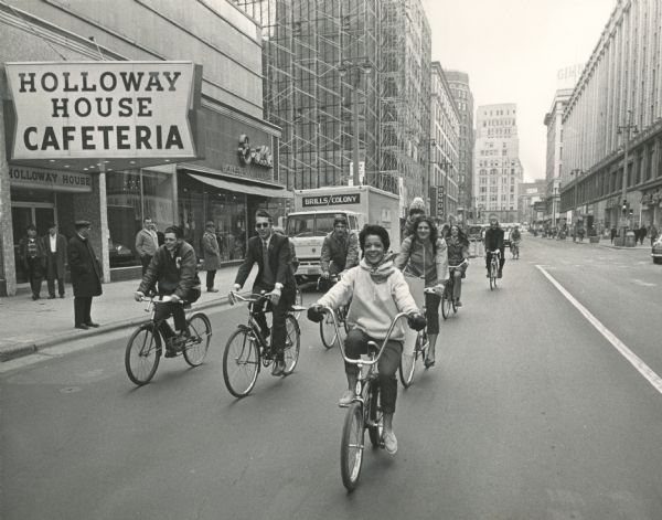 A group of bicycle riders on the street, with a smiling Vel Phillips in the lead. She is wearing a sweater, scarf, gloves, slacks and lace up shoes. Other riders are wearing suits, pants, coats and jackets. One woman has a toddler in a child seat behind her. They are just passing the Holloway House Cafeteria, a Brills Hickey-Freeman Clothes store and a Brills/Colony truck. Pedestrians are watching from the sidewalk. Tall buildings are in the background. A Gimbels sign is in the upper right corner.