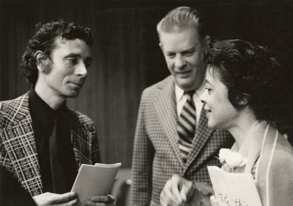 Vel Phillips chats with two men. She is wearing a dress, sweater and scarf and is holding papers and roses in one arm. The men are wearing plaid suits and neckties. The man on the left is holding some papers.