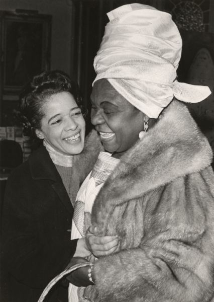 Vel Phillips is embracing Angie Brooks, former United Nations representative to Liberia. Ms. Brooks is wearing a fur coat, turban and earrings. Both women are smiling broadly. Vel is wearing an overcoat over a sweater and scarf.