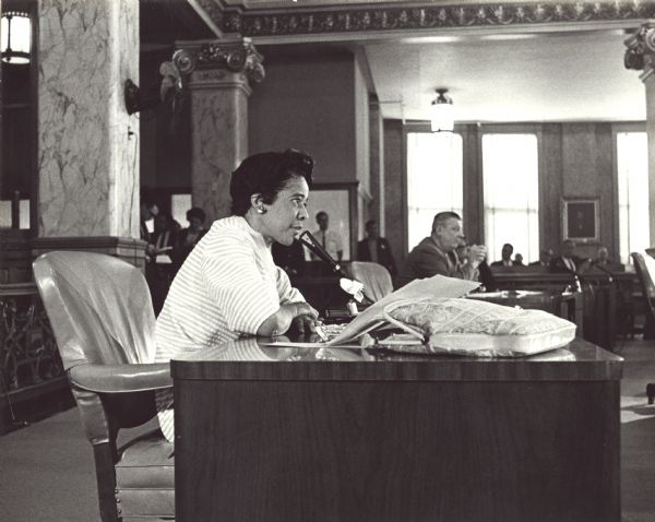 Vel Phillips speaking into a microphone at her desk in the Milwaukee Common Council. Other desks and men are behind her. The council room has ornate interior decorations.