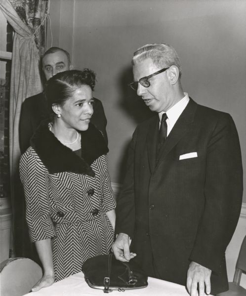 Vel Phillips stands while speaking with Arthur Goldberg, former Secretary of Labor and, at the time of the photograph, Supreme Court Justice. Another man can be seen just behind her. The men are wearing suits and neckties. Vel is wearing a herringbone suit with a dark collar. Her handbag is on the table.