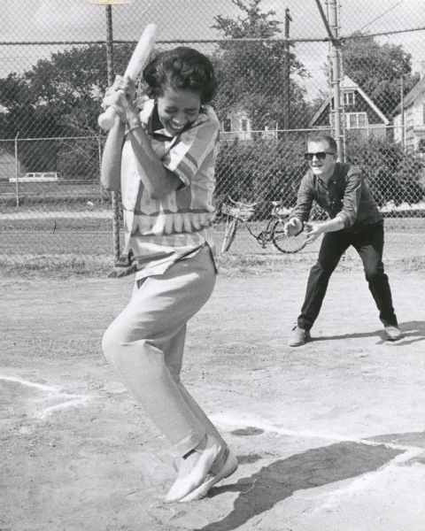 Vel Phillips grips the bat and ducks as the ball is pitched at a baseball game. The catcher is on the right. Chain link fencing, a bicycle and a residential neighborhood are in the background.