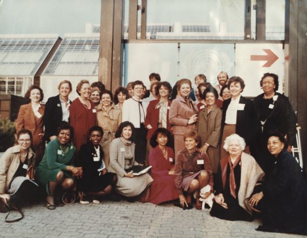 Vel Phillips in a large group portrait of mostly women. The women are wearing dresses and coats and have name tags on their lapels. A building with large glass windows is visible through the window behind them. To the left of the red arrow is the symbol for the United Nations.
