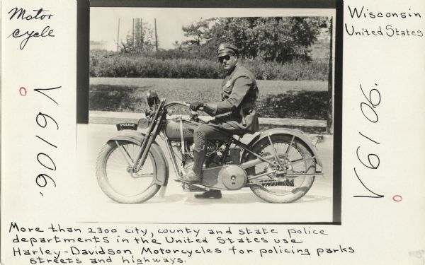 A police officer poses on the street, seated on his Harley-Davidson motorcycle. He is dressed in his uniform with police badges on his jacket and hat. A sidewalk, lawn, shrubs and trees can be seen in the background. Handwritten on three sides of the image, "Motor cycle," "V6106," "Wisconsin, United States," "V6106," "More than 2300 city, county and state police departments in the United States use Harley-Davidson Motorcycles for policing parks, streets and highways."
