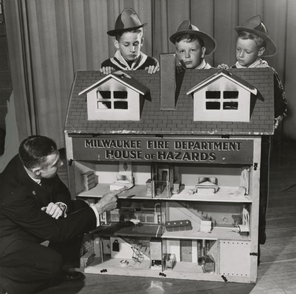 Fire hazards are pointed out in a model house by Battalion Chief Edward McCabe for Cub Scouts of Pack 566 at Manitoba School. The ten-year-old boy scouts peering over the roof are (from left): Craig Byers, William Watson and James Zamjahn. The boys are wearing their scout uniforms and fire helmets. Chief McCabe is also wearing a uniform. A sign on the model house reads "Milwaukee Fire Department House of Hazards."