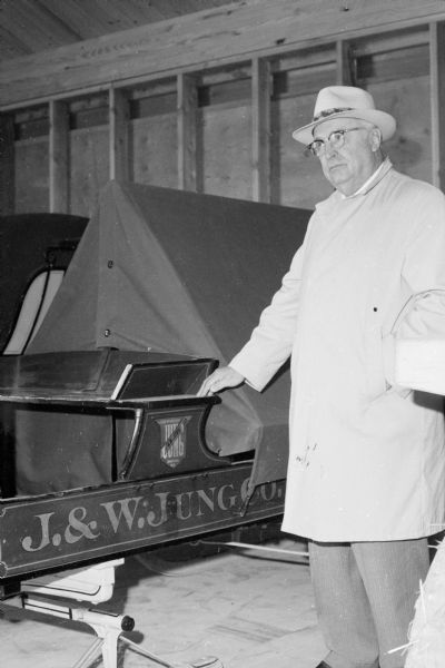 Wesley W. Jung at the Jung Carriage Museum. Mr. Jung is standing with one hand on a sleigh. On the side of the box the lettering, "J. & W. Jung Co." appears and above is the J. & W. Jung Company logo. The sled appears to have a tent pitched in the box. Mr. Jung is wearing an overcoat, hat and glasses.