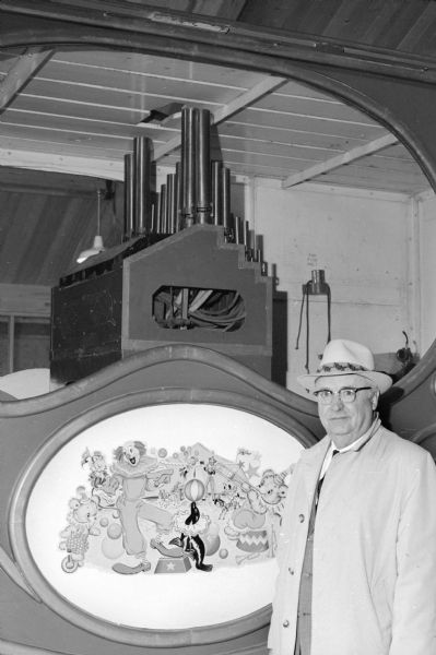 Wesley W. Jung at the Jung Carriage Museum. Mr. Jung is standing on the right in front of a circus wagon. Painted inside of an oval on the side is a circus themed illustration that includes a clown, bears, a monkey, a seal and a tent. A calliope can be seen inside the wagon. Mr. Jung is wearing an overcoat, hat and glasses.