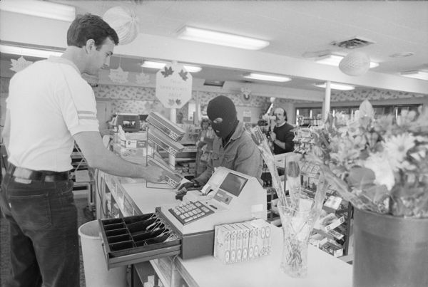 This image was captured during the re-enactment of a crime that took place at a convenience store on Tuesday, April 22nd, 1986. The killing of Andrew N. Nehmer was re-enacted as police believe it happened for the filming of a Crime Stoppers public service message for the Madison Police Department. Police Officer Jay Lengfeld, white shirt, depicted Nehmer, and Detective Rick Miller portrayed the robber. In the re-enactment, Lengfeld takes money out of the cash register as demanded by Miller. Filming was done by Dave Friend.