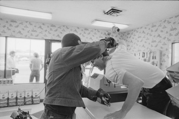 This image was captured during the re-enactment of a crime that took place at a convenience store on Tuesday, April 22nd, 1986. The killing of Andrew N. Nehmer was re-enacted as police believe it happened for the filming of a Crime Stoppers public service message for the Madison Police Department. Police Officer Jay Lengfeld, white shirt, depicted Nehmer, and Detective Rick Miller portrayed the robber. In the re-enactment, Miller portrays the stabbing of Nehmer. Filming was done by Dave Friend. Two men can be seen standing outside the window near the entrance.