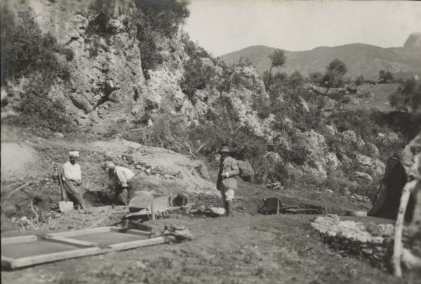 Ensemble of diggings at Ali Bacha. View looking downhill towards three men working on an archaeological excavation in Algeria, Africa. On the left an indigenous man is leaning back against the side of the trench with his wrists crossed. The indigenous man in the center is digging with a pick. Both men are wearing work clothes and head wraps. A third man down in the trench is bending over. The man standing on the right may be M. Debruge, who is wearing European clothing. Two wheelbarrows and other equipment are on the site, and a tent is partially visible on the right. Rock formations, trees and shrubs surround the site and rocky hills are in the background.
