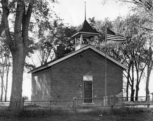 Dunham School, standing in a grove of trees, surrounded by a fence. The building is built of brick and has a bell tower and chimney, with a front door with a transom window in the center. The windows on the left side of the building are bricked over and repairs to the corner bricks can be seen. An American flag flies on a flagpole in front of the entrance.