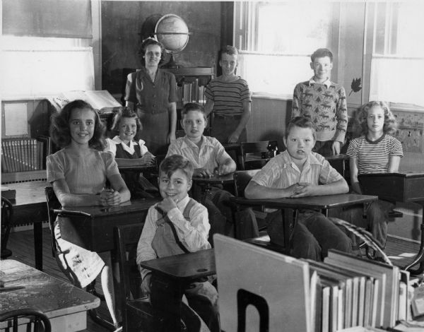 Students and teacher in the classroom at Dunham School. The room is filled with desks, chairs, books, a blackboard and a globe. Three large window can be seen in the background. Identified are, in foreground: Peter Behrens, and behind him, left to right: Mary Balogh, Marleen Looker, Earl Richard Quinney, Ralph Quinney, Betty Gies. Standing in the back, left to right, teacher Evelyn Olsen, Lawrence Duesterbeck and Patrick McDonald.