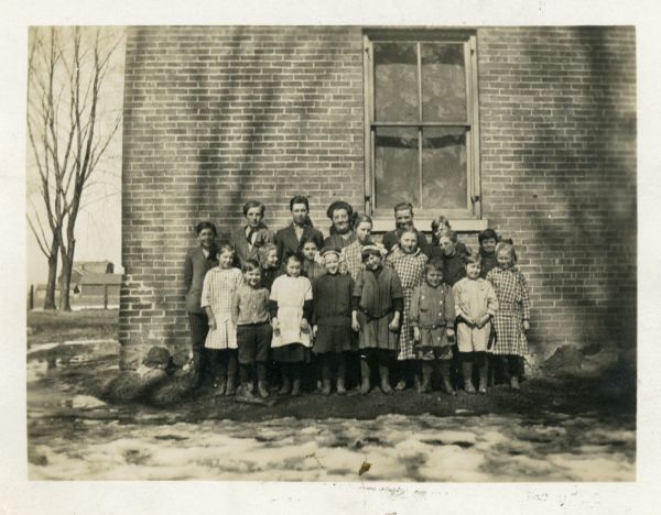 The students of Dunham School pose outdoors with the school building in the background. Identified are (front, left to right) LaVerne Campbell, Mildred Peterson, Mary Jones, Lester Peterson, Helen Dutcher, (middle row, left to right) Iva Dutcher, Helen Peterson, Blanche Macafee, Clara Peterson, Glenn Peterson, Roy Macafee, Alice Kittleson, (back row, left to right) August Schultz, Olaf Peterson, Floyd Quinney, teacher Lillian Wilear, Walter Duesterbeck, Bert Duesterbeck. The teacher and girls are wearing dresses and the boys are wearing trousers and shirts. The boys in the back row are also wearing coats. Everyone is wearing stockings and shoes. Snow can be seen in the foreground.