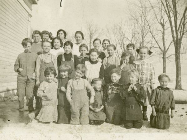 An outdoor group portrait of the students of South Heart Prairie School. Identified on the back of the portrait are Eda Papcke, Ella Koestler, Ruth Gutzmer, Ellen Gutzmer, Alice Jordan, Alice Holloway (first on the right in the second row), Rosie Bazen, Ollie Bazen, Edna Bazen, Irena Bazen, Hazel McQuillin, Leo McQuillin, Neil Bogie, Roy Kitzman, Harry Kitzman, Walter Gutzmer, Isabella Butler, Vera Butler, Robert Dunham, Wave Henry, Loren Henry, Frank Henry and Fred Henry. The girls are wearing dresses and the boys are wearing trousers, bib overalls, shirts and coats. The side of the schoolhouse can be seen on the left. There is snow on the ground and the trees are leafless.