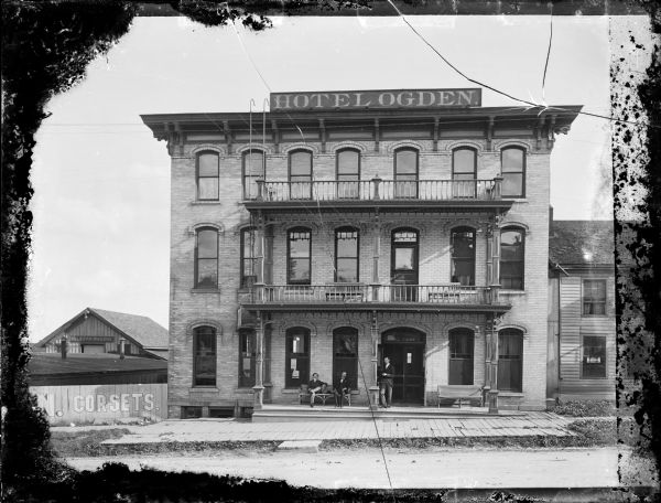 Exterior view of the front of the Hotel Ogden located at 149 East Wilson Street. Three men wearing suits are relaxing on the porch, with two men sitting on a bench and one man standing. The hotel facade has two balconies above the porch, 7 windows on each of the three stories, and a fire escape. The fence on the left is painted with an advertising sign that reads: "Corsets." In the background is a building with a sign above the doorway that reads: "Milburn Wagons."