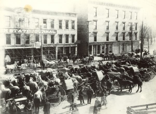 Elevated view of the farmer's market on East Washington Avenue near storefronts. Many horse-drawn buggies and wagons are lined up on both sides of the avenue. Buyers and vendors are walking, standing or sitting on the sidewalk and in the street. Sign on storefronts are: "[Wisconsi?]n State Journal," "Piper Brothers Grocers & Seeds," "Conklin & Sons, Coal, Wood and Ice," "J.P. Halbach, Harness & Saddlery" and "Hides Bought Here."
