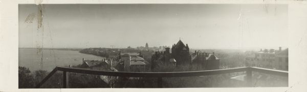 Elevated panoramic view of the isthmus from campus, looking east. The location of the photographer is unknown, but a railing is in the foreground. The largest building in the center is  Science Hall. Lake Mendota is on the left, Lake Monona is in the far distance on the right. The Wisconsin State Capitol dome is silhouetted in the background.
