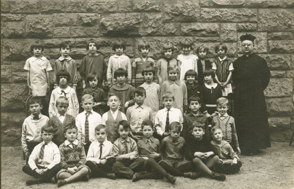 Students pose outdoors in front of a stone wall at Saint Raphael's School. David O'Dea is seated cross-legged on the ground, 3rd from the right. He is possibly in the 3rd or 4th grade. The priest is Father Murphy.