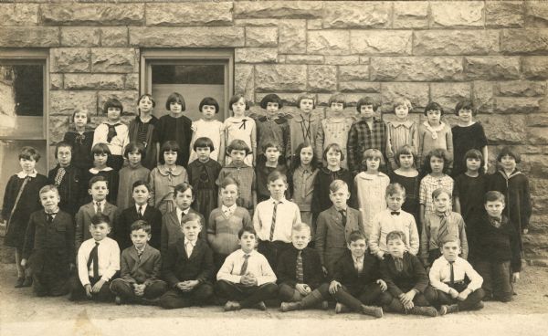 Students pose outdoors in front of a stone building at Saint Raphael's School. Mary Julia O'Dea (born 1917) is standing in the third row, second from the right. She is possibly in the 4th grade. Two large windows are behind the students.